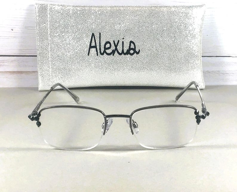 May include: Rimless eyeglasses with a silver frame and decorative black accents at the temples. The glasses are in front of a white, textured eyeglass case with the name "Alexia" in black script.