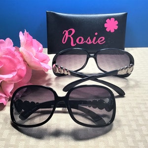 Puede incluir: Dos pares de gafas de sol negras con detalles plateados en las monturas. Las gafas de sol est&aacute;n sobre una superficie beige con una funda de gafas de sol negra con la palabra "Rosie" en letras rosas.