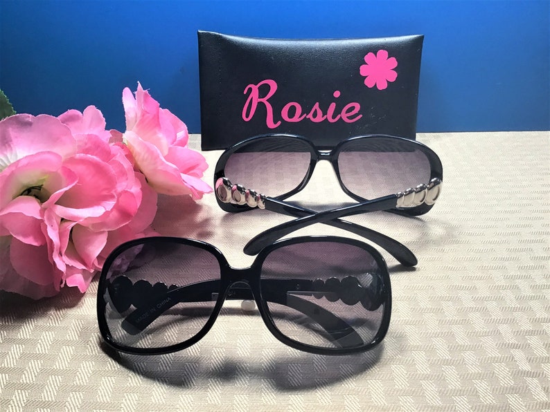 Puede incluir: Dos pares de gafas de sol negras con detalles plateados en las monturas. Las gafas de sol est&aacute;n sobre una superficie beige con una funda de gafas de sol negra con la palabra "Rosie" en letras rosas.