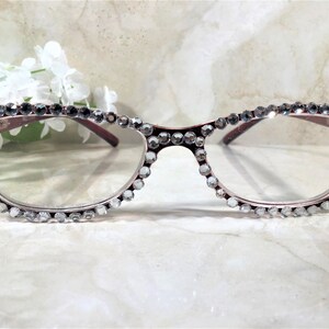 May include: A pair of burgundy-framed eyeglasses with clear lenses. The frames are embellished with numerous clear, sparkling rhinestones. The glasses have a cat-eye shape and are set against a soft, blurred background with white flowers.