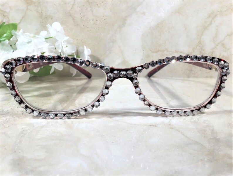 May include: A pair of burgundy-framed eyeglasses with clear lenses. The frames are embellished with numerous clear, sparkling rhinestones. The glasses have a cat-eye shape and are set against a soft, blurred background with white flowers.