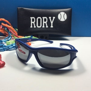 Puede incluir: Gafas de sol azules con lentes espejados y una funda negra con el nombre "RORY" y un gr&aacute;fico de b&eacute;isbol blanco.