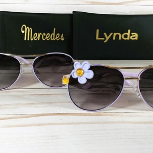 Pu&ograve; includere: Due paia di occhiali da sole aviator viola con dettagli dorati. Un paio ha una stella dorata sulla lente sinistra e l'altro ha un fiore viola con un centro giallo sulla lente destra. Gli occhiali da sole sono su una superficie di legno bianco con due custodie nere dietro di loro. Le custodie hanno i nomi "Mercedes" e "Lynda" scritti in lettere dorate.