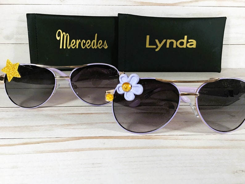 Pu&ograve; includere: Due paia di occhiali da sole aviator viola con dettagli dorati. Un paio ha una stella dorata sulla lente sinistra e l'altro ha un fiore viola con un centro giallo sulla lente destra. Gli occhiali da sole sono su una superficie di legno bianco con due custodie nere dietro di loro. Le custodie hanno i nomi "Mercedes" e "Lynda" scritti in lettere dorate.