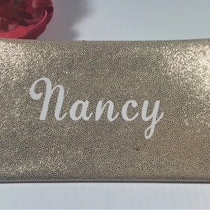 Pu&ograve; includere: Una pochette rettangolare dorata con una superficie strutturata. Il nome "Nancy" &egrave; scritto in bianco sulla parte anteriore. Sullo sfondo &egrave; visibile un fiore rosso.