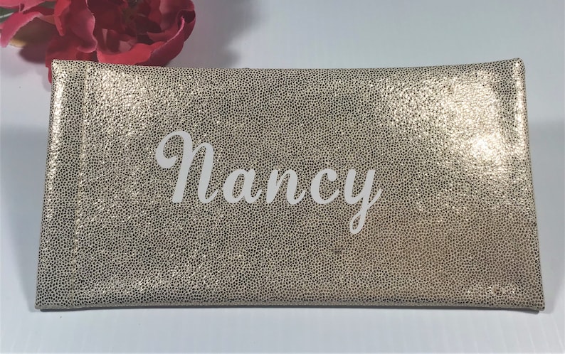 Pu&ograve; includere: Una pochette rettangolare dorata con una superficie strutturata. Il nome "Nancy" &egrave; scritto in bianco sulla parte anteriore. Sullo sfondo &egrave; visibile un fiore rosso.