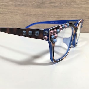 May include: Blue and brown eyeglasses with a leopard print pattern on the frames. The frames are decorated with blue and pink rhinestones. The glasses have clear lenses.