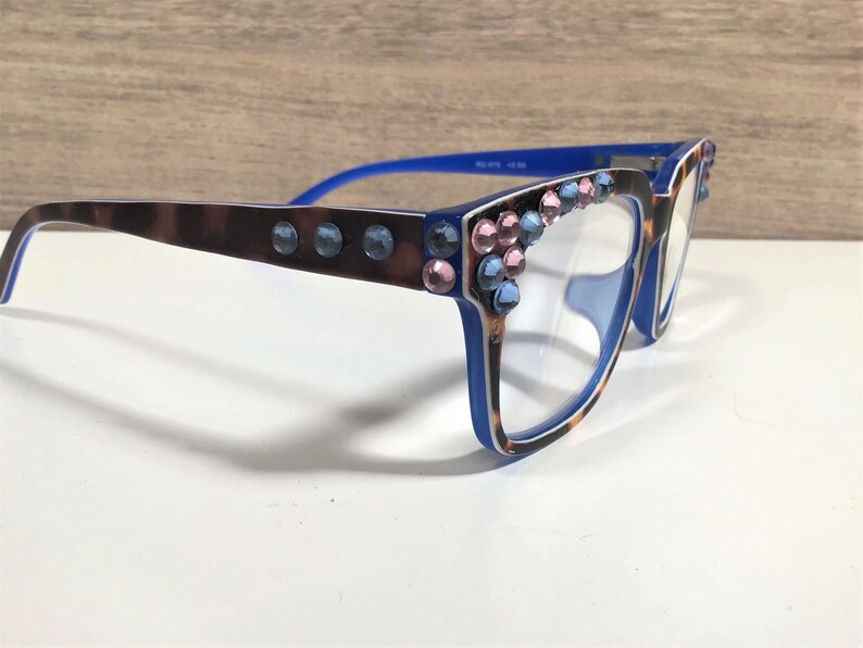 May include: Blue and brown eyeglasses with a leopard print pattern on the frames. The frames are decorated with blue and pink rhinestones. The glasses have clear lenses.