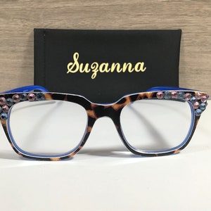 May include: Tortoiseshell patterned eyeglasses with blue detailing and pink and blue rhinestone embellishments. A black case with the name "Suzanna" in gold script is behind the glasses.