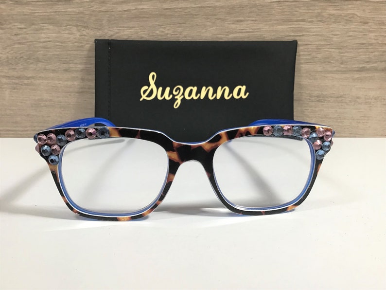 May include: Tortoiseshell patterned eyeglasses with blue detailing and pink and blue rhinestone embellishments. A black case with the name "Suzanna" in gold script is behind the glasses.