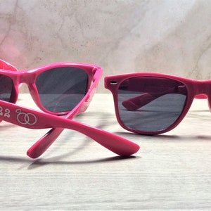May include: Two pairs of pink sunglasses with black lenses. One pair has the text "July '22" and two interlocking rings on the side.
