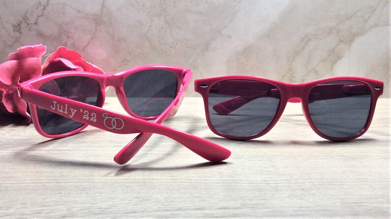 May include: Two pairs of pink sunglasses with black lenses. One pair has the text "July '22" and two interlocking rings on the side.