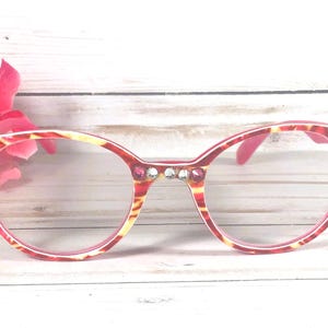 May include: Eyeglasses with a red and yellow tortoiseshell pattern frame, featuring clear lenses and decorative rhinestones embedded along the bridge and outer corners of the frame front, providing a detailed and ornate appearance.