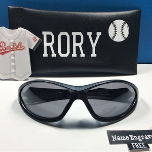 May include: Black sunglasses with a textured frame and gray lenses. The sunglasses come with a black case that has the name "RORY" and a baseball graphic printed on it. The case also has a baseball jersey graphic with the word "Baseball" printed on it. The text "Name Engraved FREE" is printed on a white label.