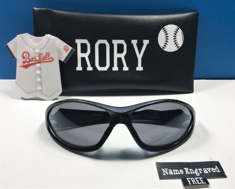 May include: Black sunglasses with a textured frame and gray lenses. The sunglasses come with a black case that has the name "RORY" and a baseball graphic printed on it. The case also has a baseball jersey graphic with the word "Baseball" printed on it. The text "Name Engraved FREE" is printed on a white label.