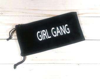 May include: A black rectangular soft pouch with a drawstring closure on the left side. The text GIRL GANG is printed in white capital letters across the center of the pouch. The pouch has a smooth, matte finish and a simple, functional design.