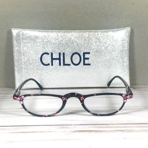 Pu&ograve; includere: Occhiali da lettura neri con un motivo floreale rosa sulle montature e piccoli accenti rosa agli angoli. Gli occhiali sono posizionati davanti a una custodia rettangolare glitterata argento con la parola CHLOE stampata in lettere blu scuro.