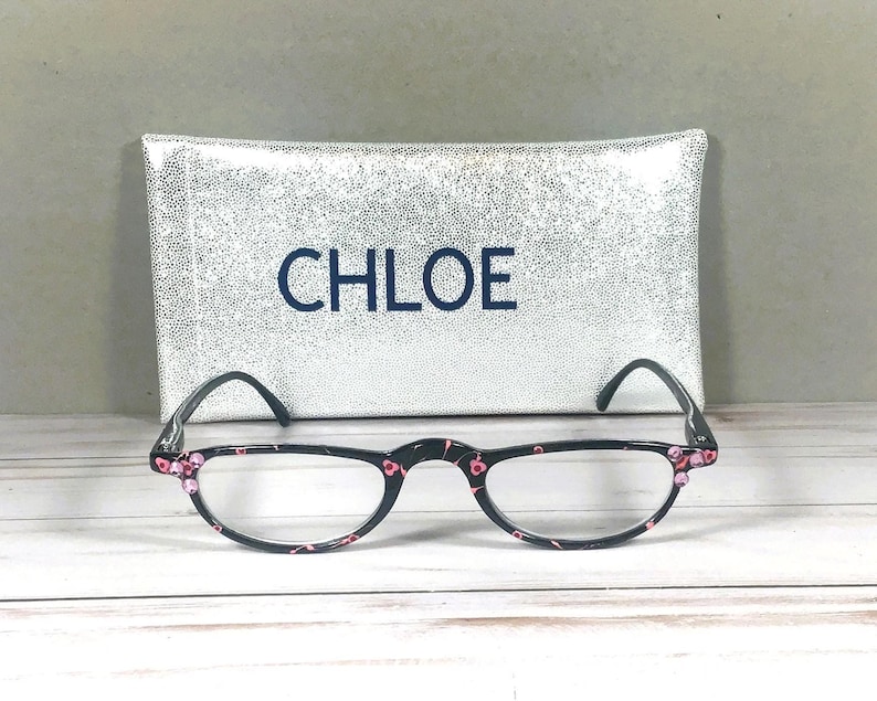 Pu&ograve; includere: Occhiali da lettura neri con un motivo floreale rosa sulle montature e piccoli accenti rosa agli angoli. Gli occhiali sono posizionati davanti a una custodia rettangolare glitterata argento con la parola CHLOE stampata in lettere blu scuro.