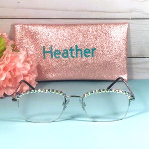 May include: Silver-rimmed glasses featuring a row of iridescent rhinestones along the top frame. Behind the glasses sits a rectangular rose gold glitter case with the name Heather printed in teal letters on the front surface.