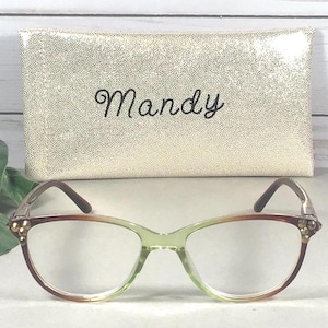 Pu&ograve; includere: Occhiali con montatura sfumata verde e marrone e custodia dorata. La custodia &egrave; strutturata e riporta il nome "Mandy" in corsivo. Gli occhiali hanno una forma a occhi di gatto con dettagli decorativi.