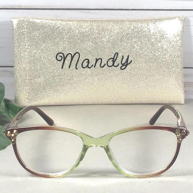 Pu&ograve; includere: Occhiali con montatura sfumata verde e marrone e custodia dorata. La custodia &egrave; strutturata e riporta il nome "Mandy" in corsivo. Gli occhiali hanno una forma a occhi di gatto con dettagli decorativi.