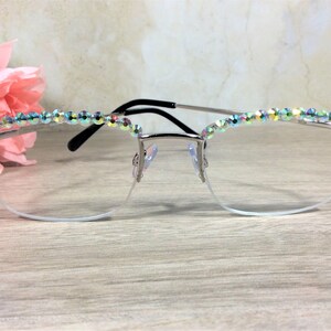 May include: Silver-toned semi-rimless eyeglasses featuring a row of iridescent, multi-colored crystals embedded along the top of the frames. The glasses have clear lenses, a thin metal bridge, and black temple tips for a secure fit.