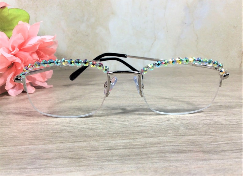 May include: Silver-toned semi-rimless eyeglasses featuring a row of iridescent, multi-colored crystals embedded along the top of the frames. The glasses have clear lenses, a thin metal bridge, and black temple tips for a secure fit.