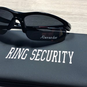 May include: Black sunglasses with a gold tint on the lenses and a black case with the text "RING SECURITY" printed in white.