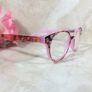May include: Pink and tortoiseshell eyeglasses with clear lenses. The frames are embellished with small, sparkling purple rhinestones. The temples have a copper-colored metallic finish. A pink flower is in the background.