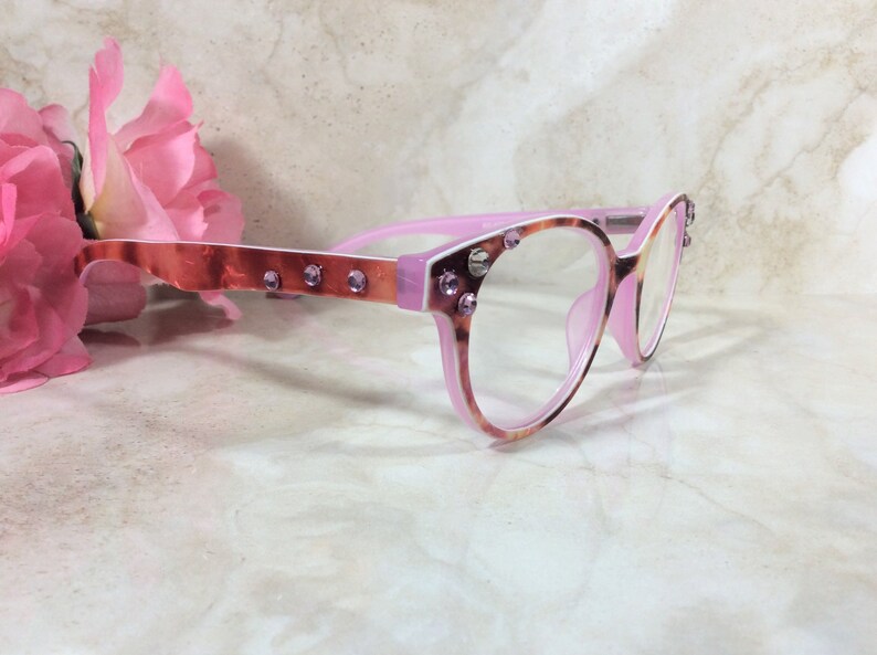 May include: Pink and tortoiseshell eyeglasses with clear lenses. The frames are embellished with small, sparkling purple rhinestones. The temples have a copper-colored metallic finish. A pink flower is in the background.