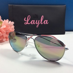 May include: Silver aviator sunglasses with a pink frame and iridescent lenses. The sunglasses are in a black case with the name "Layla" printed on it in pink.
