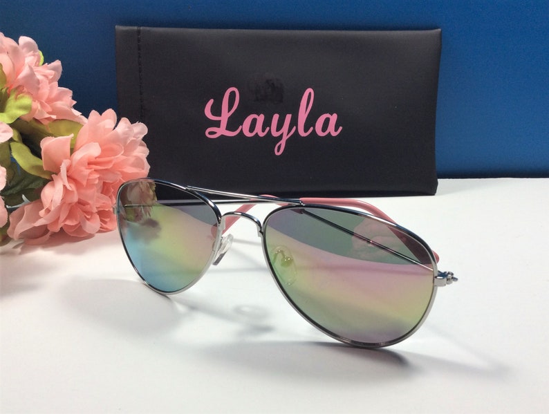 May include: Silver aviator sunglasses with a pink frame and iridescent lenses. The sunglasses are in a black case with the name "Layla" printed on it in pink.