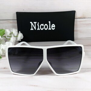 K&ouml;nnte beinhalten: Wei&szlig;e quadratische Sonnenbrille mit dunkelgrauen Gl&auml;sern. Die Sonnenbrille liegt auf einer wei&szlig;en Holzoberfl&auml;che. Die Sonnenbrille wird mit einem schwarzen Etui geliefert, auf dem das Wort "Nicole" in Wei&szlig; aufgedruckt ist.