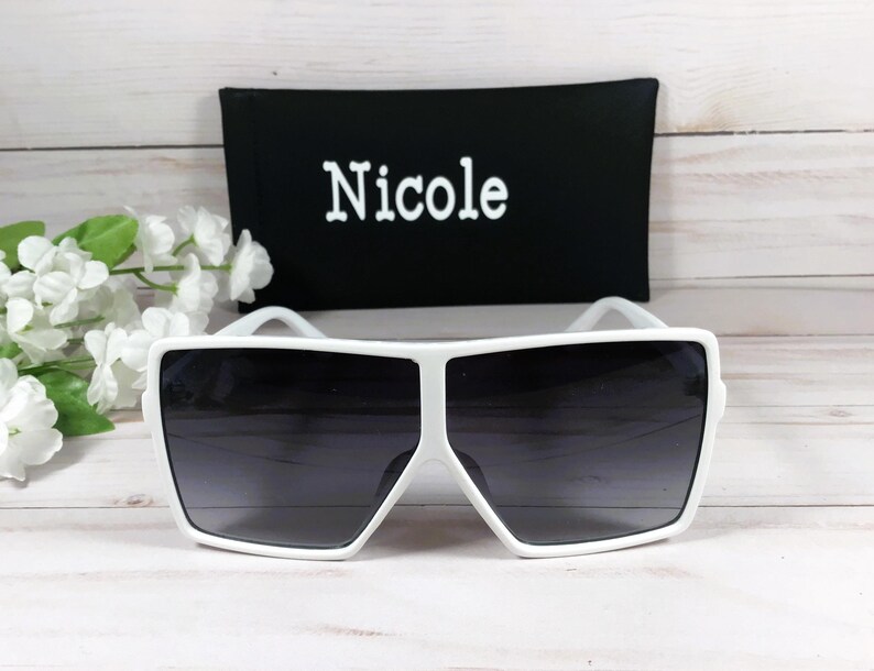 K&ouml;nnte beinhalten: Wei&szlig;e quadratische Sonnenbrille mit dunkelgrauen Gl&auml;sern. Die Sonnenbrille liegt auf einer wei&szlig;en Holzoberfl&auml;che. Die Sonnenbrille wird mit einem schwarzen Etui geliefert, auf dem das Wort "Nicole" in Wei&szlig; aufgedruckt ist.