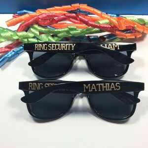 Op de afbeelding: Twee paar zwarte zonnebrillen met gouden letters. Het bovenste paar zegt "RING SECURITY" en "LIAM". Het onderste paar zegt "RING SECURITY" en "MATHIAS".
