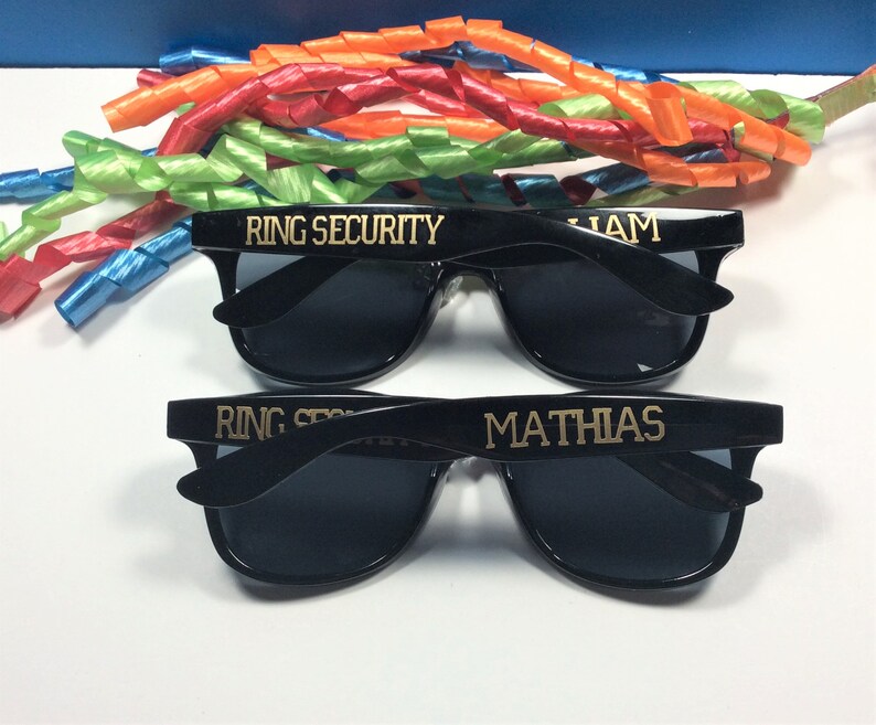 Op de afbeelding: Twee paar zwarte zonnebrillen met gouden letters. Het bovenste paar zegt "RING SECURITY" en "LIAM". Het onderste paar zegt "RING SECURITY" en "MATHIAS".