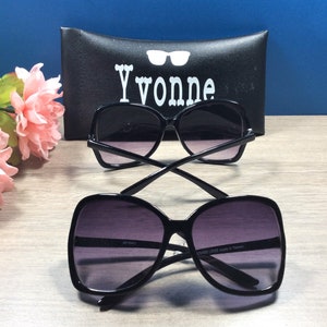 May include: Two pairs of black sunglasses with dark lenses are displayed on a wooden surface. The sunglasses have a square, oversized frame. A black case with the name "Yvonne" in white lettering is in the background. Pink flowers are on the left side.
