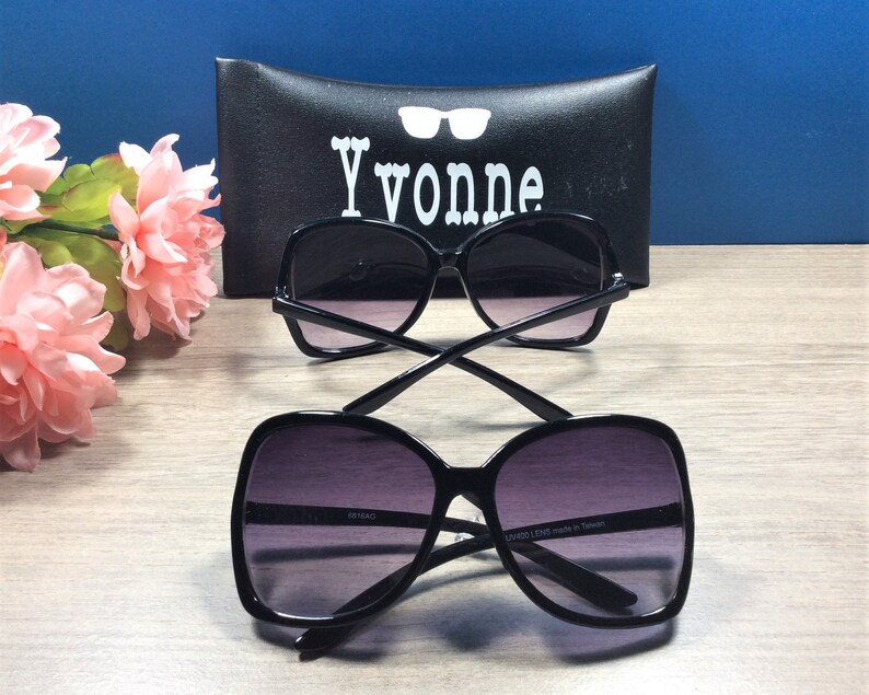 May include: Two pairs of black sunglasses with dark lenses are displayed on a wooden surface. The sunglasses have a square, oversized frame. A black case with the name "Yvonne" in white lettering is in the background. Pink flowers are on the left side.
