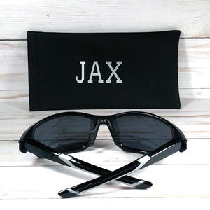 May include: Black sunglasses with dark lenses and a black case. The case has the name "JAX" printed in silver. The sunglasses have a modern design with silver accents on the arms. The image is set against a white wooden background.