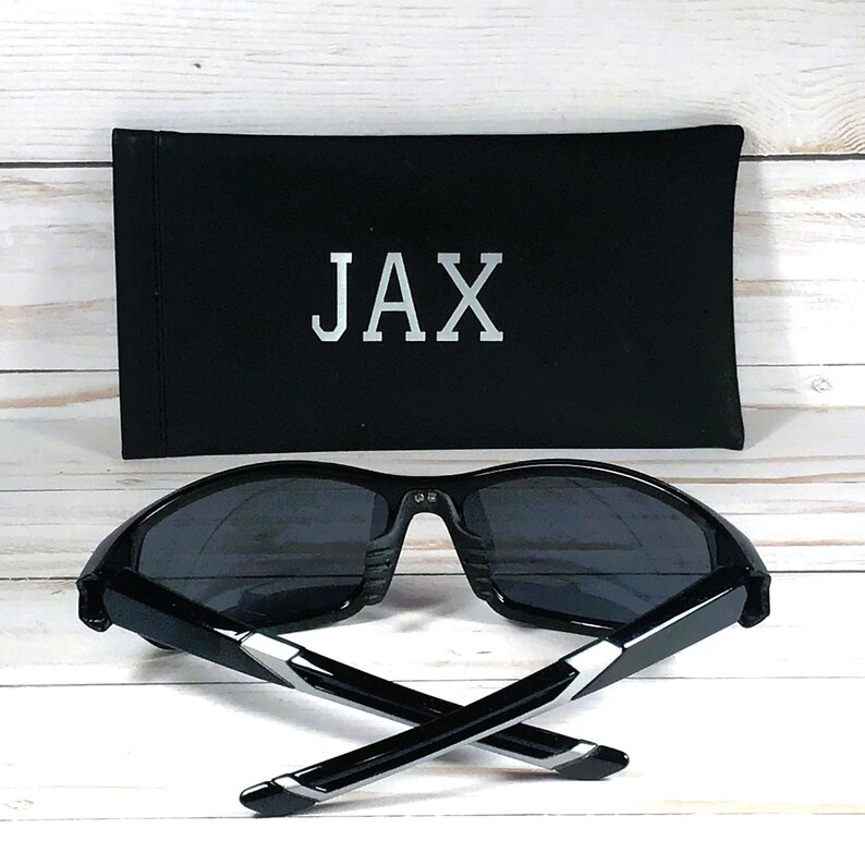 May include: Black sunglasses with dark lenses and a black case. The case has the name "JAX" printed in silver. The sunglasses have a modern design with silver accents on the arms. The image is set against a white wooden background.