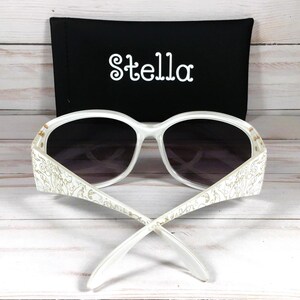 K&ouml;nnte beinhalten: Wei&szlig;e Sonnenbrille mit floralem Design an den B&uuml;geln. Die Sonnenbrille hat ein schwarzes Etui mit dem Aufdruck "Stella".