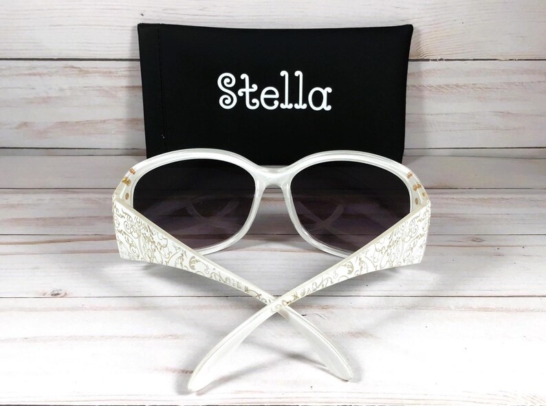 K&ouml;nnte beinhalten: Wei&szlig;e Sonnenbrille mit floralem Design an den B&uuml;geln. Die Sonnenbrille hat ein schwarzes Etui mit dem Aufdruck "Stella".