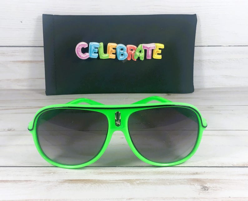 Puede incluir: Gafas de sol de aviador verde brillante con lentes oscuras. Detr&aacute;s de las gafas de sol hay un estuche negro con la palabra "CELEBRATE" en letras coloridas. Las gafas de sol tienen un dise&ntilde;o audaz y moderno.