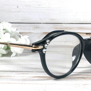 May include: Black spectacles with clear lenses featuring a gold-toned metal temple arm and three small, clear, circular rhinestones embedded on the outer corner of each frame front, providing a decorative accent to the round-shaped eyewear.