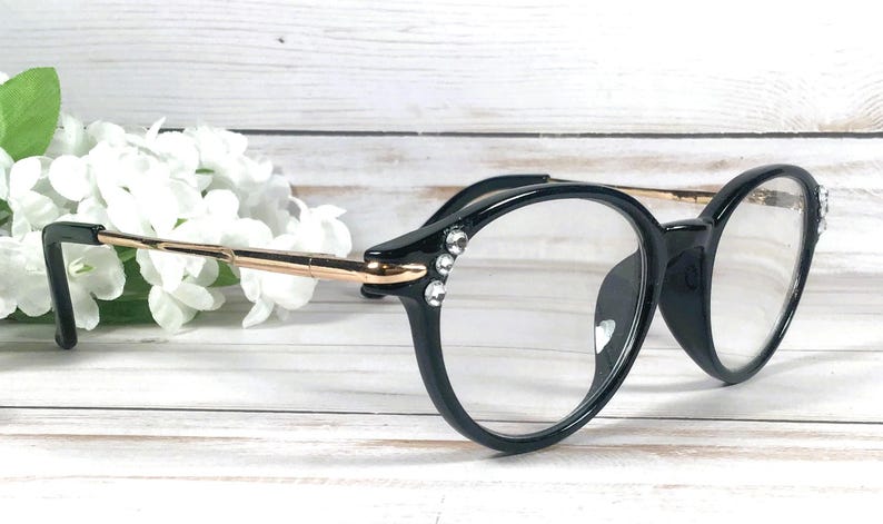 May include: Black spectacles with clear lenses featuring a gold-toned metal temple arm and three small, clear, circular rhinestones embedded on the outer corner of each frame front, providing a decorative accent to the round-shaped eyewear.
