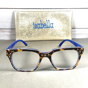 Puede incluir: Gafas con montura de carey, patillas azules y tachuelas doradas. Las gafas est&aacute;n delante de un bolso de mano dorado con el nombre "isabella" bordado en azul.