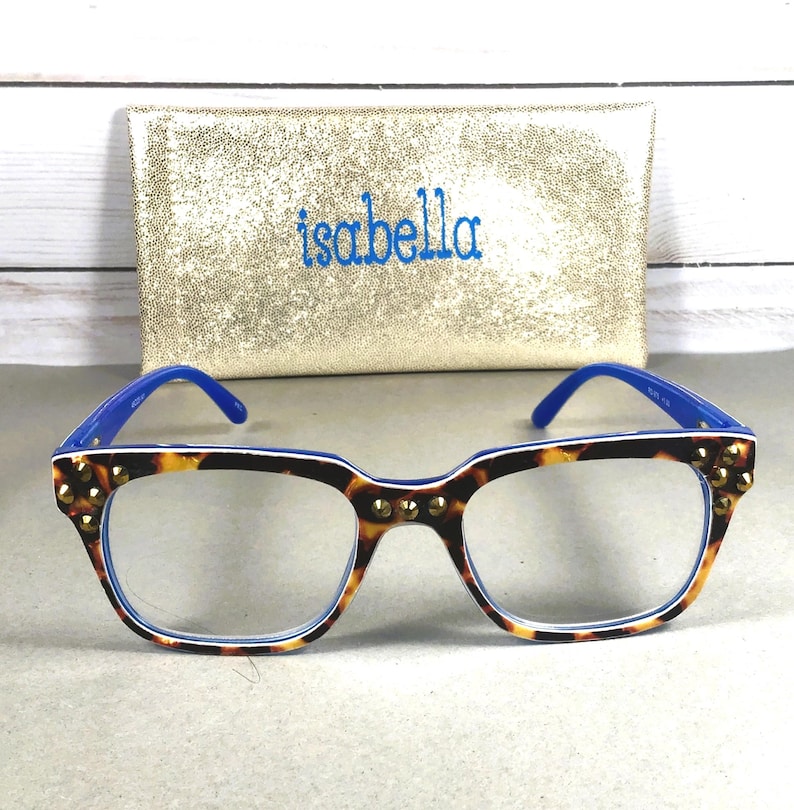 Puede incluir: Gafas con montura de carey, patillas azules y tachuelas doradas. Las gafas est&aacute;n delante de un bolso de mano dorado con el nombre "isabella" bordado en azul.