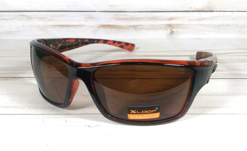 May include: Brown-tinted sunglasses with a black frame and red-brown patterned accents. The sunglasses have a rectangular shape and a curved design. The lenses are brown and the frame has a glossy finish. The brand name "XLOOP" is visible on a sticker.