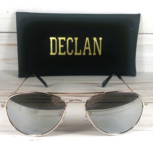 May include: Aviator sunglasses with thin gold-tone metal frames and reflective silver lenses. The glasses feature a double bridge design and black temple tips. Behind the sunglasses sits a black rectangular case with the word DECLAN in gold.