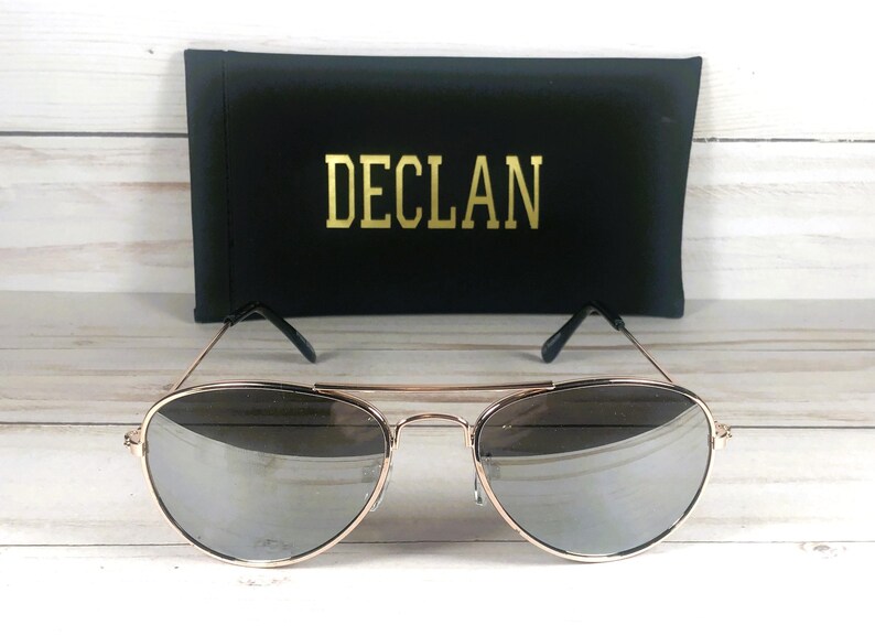 May include: Aviator sunglasses with thin gold-tone metal frames and reflective silver lenses. The glasses feature a double bridge design and black temple tips. Behind the sunglasses sits a black rectangular case with the word DECLAN in gold.